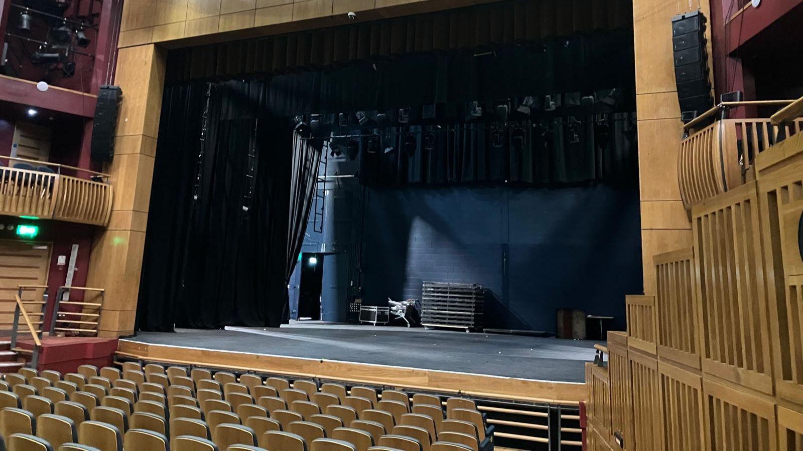 A theatre stage with seats in front made of a light coloured wood. Balconies on wither side of the stage in the same wood. 