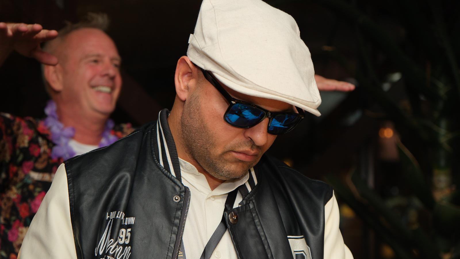 A man wearing a white flat cap, sunglasses, a white shirt and a black and white US college-style letterman jacket, DJs. Norman Cook stands in the background watching.