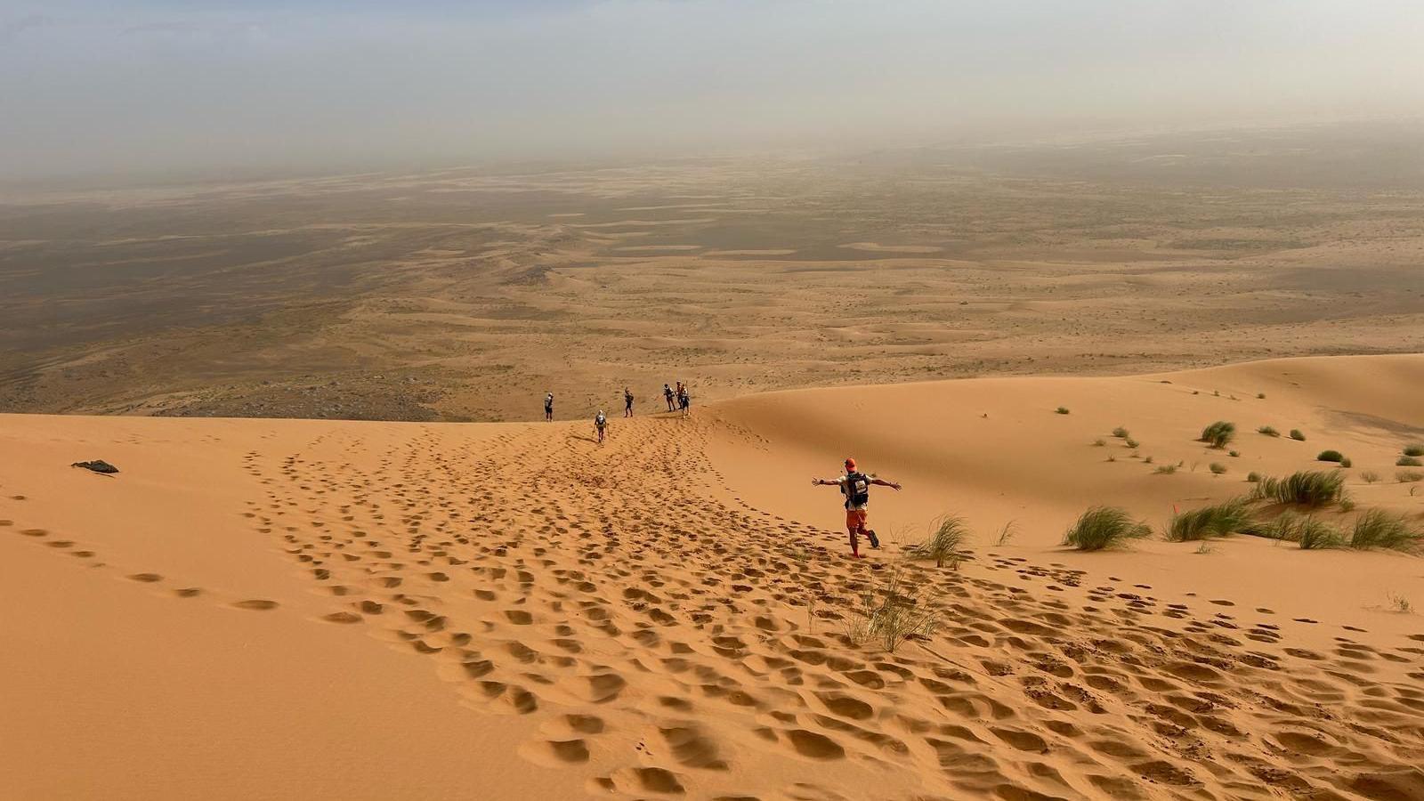 A wide expanse of orange coloured sand with a man wearing a cap running with his arms open wide and five other runners in the distance.