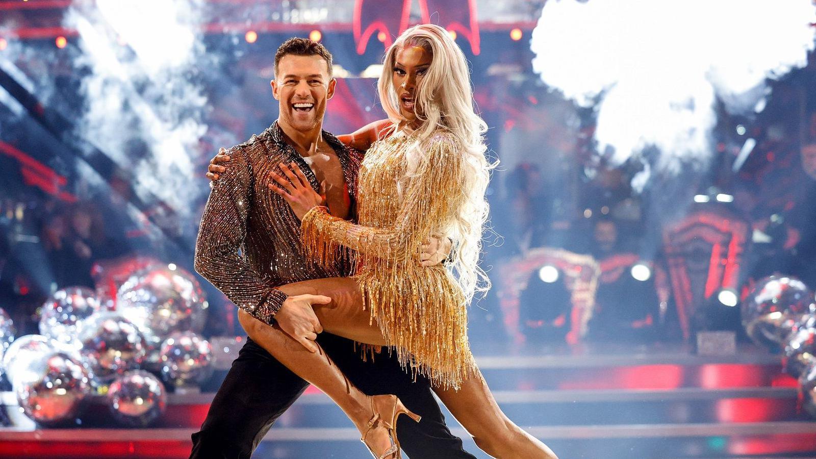 Tayce dancing with Kai Widdrington on Strictly. Tayce is wearing a short gold fitted dress and has long blonde hair. Kai is wearing a sheer black and silver shirt and black trousers and there is a Christmas tree in the background