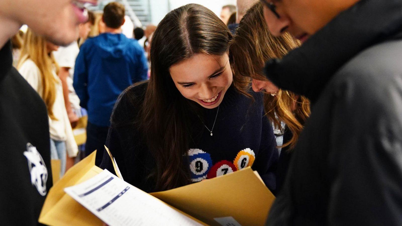 A group of pupils open their exam results. Directly in front of the camera is a female student with long straight brown hair, who wears a smiler necklace and a navy blue jumper with embroidered snooker balls. She is smiling while looking at her results with her friend, who stands to her right and whose face is obscured by her long wavy hair.