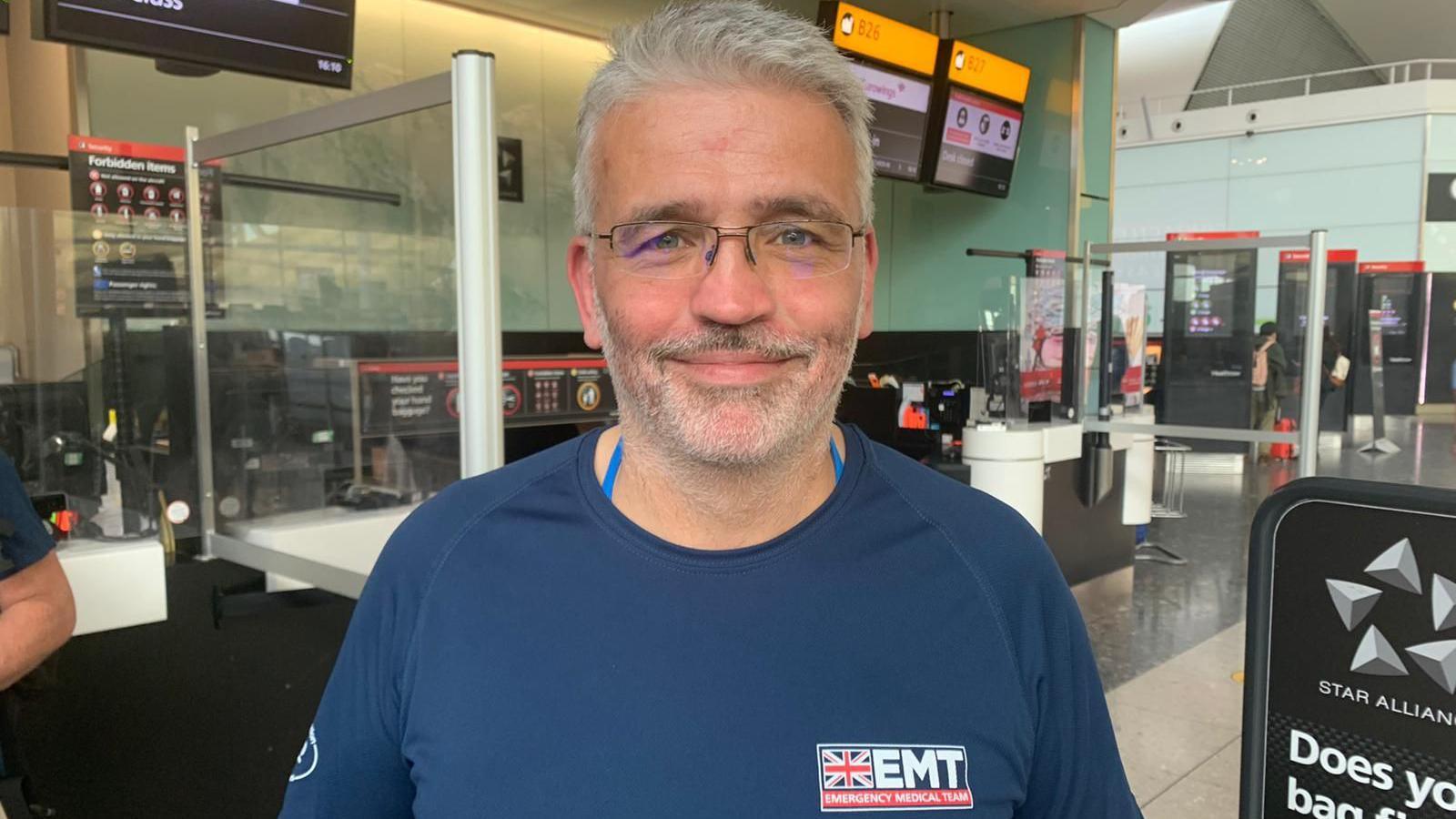 A man, David Anderson, looks into the camera. He has grey hair, blue eyes and glasses and is smiling kindly into the camera. He stands in an airport. 