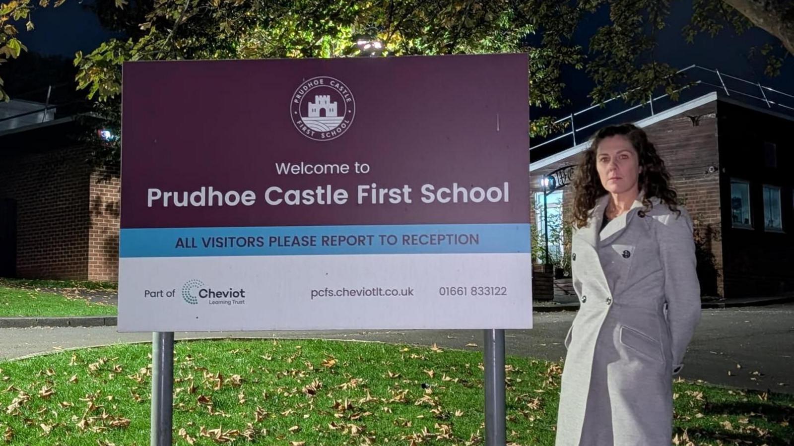 Stacey Martin wearing a grey trench coat and standing by a large purple sign which says "Prudhoe Castle First School". Ms Martin has black hair and is looking at the camera. There is green grass below littered with brown leaves. There is a school building in the background. It is dark.