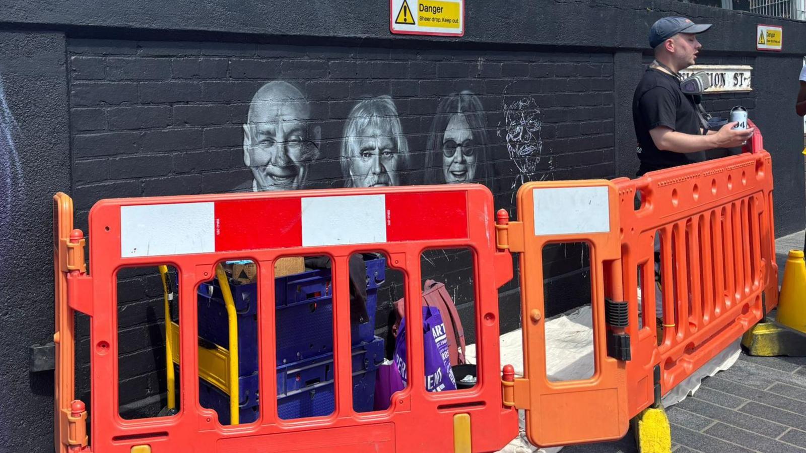 A mural in progress of Black Sabbath. It is gated off by orange fences and a the artist is stood talking to people