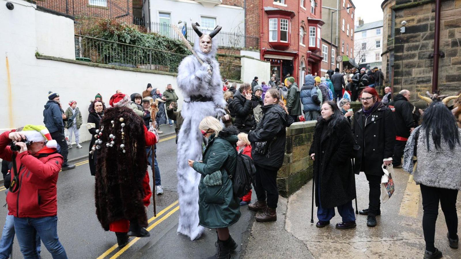 A crowd of people watch performances in a street by a person dressed in a white fur suit with horns and white face paint.