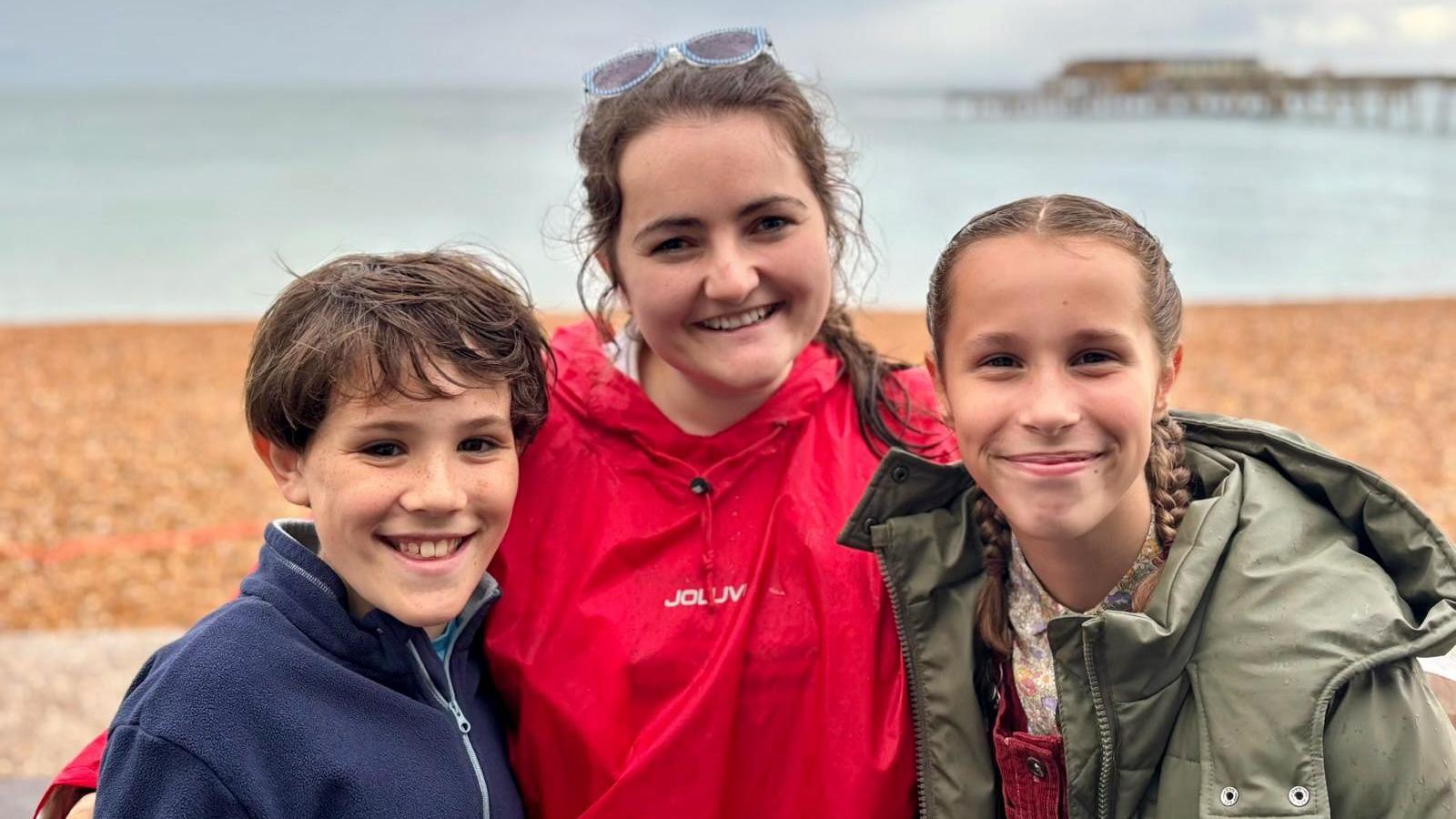 A woman smiles as she stands with a boy and a girl on a beach. Behind them is the beach, the sea and a pier.