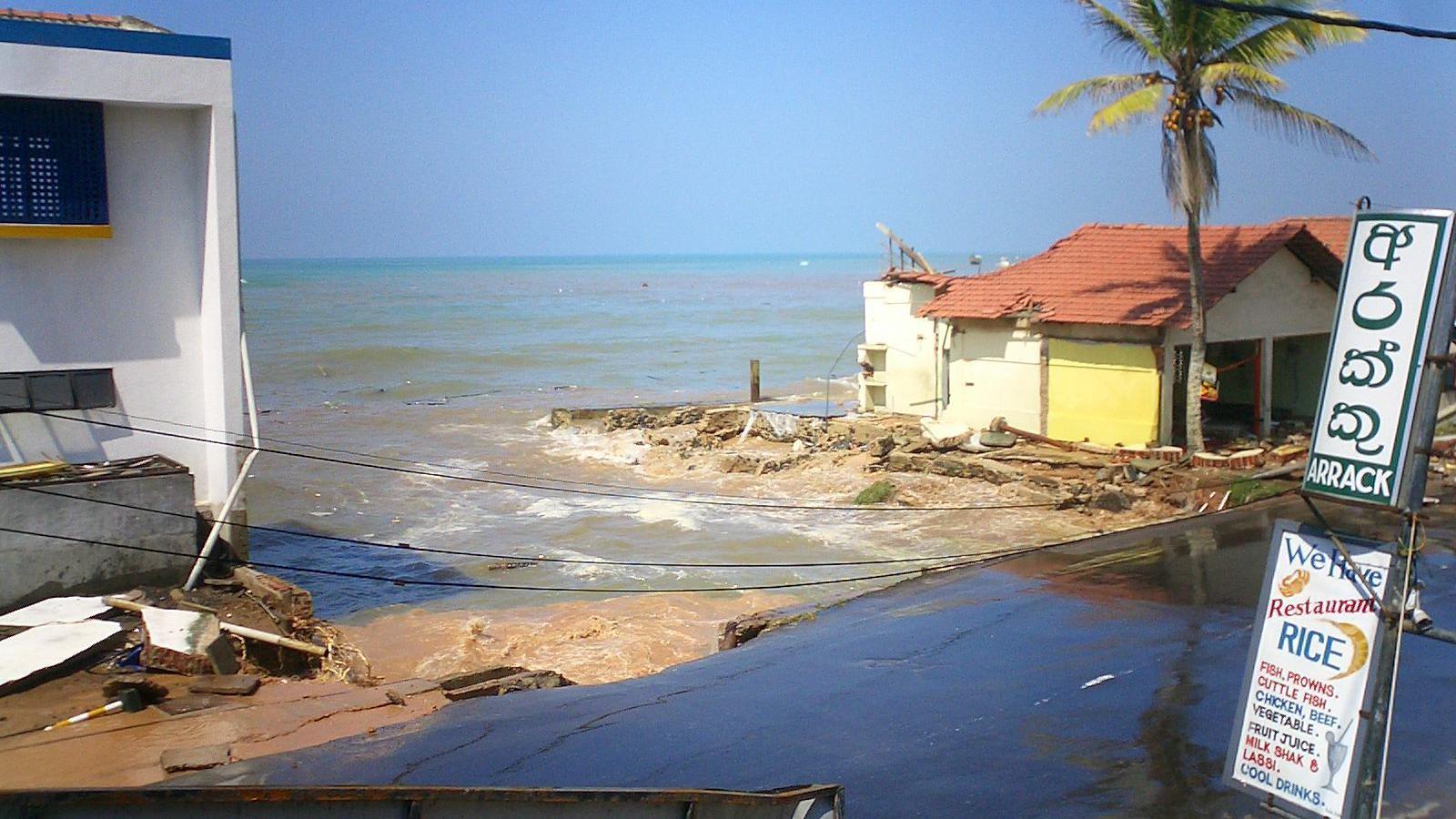 A small one-storey building, partially wrecked, stands near the sea by a flooded street.