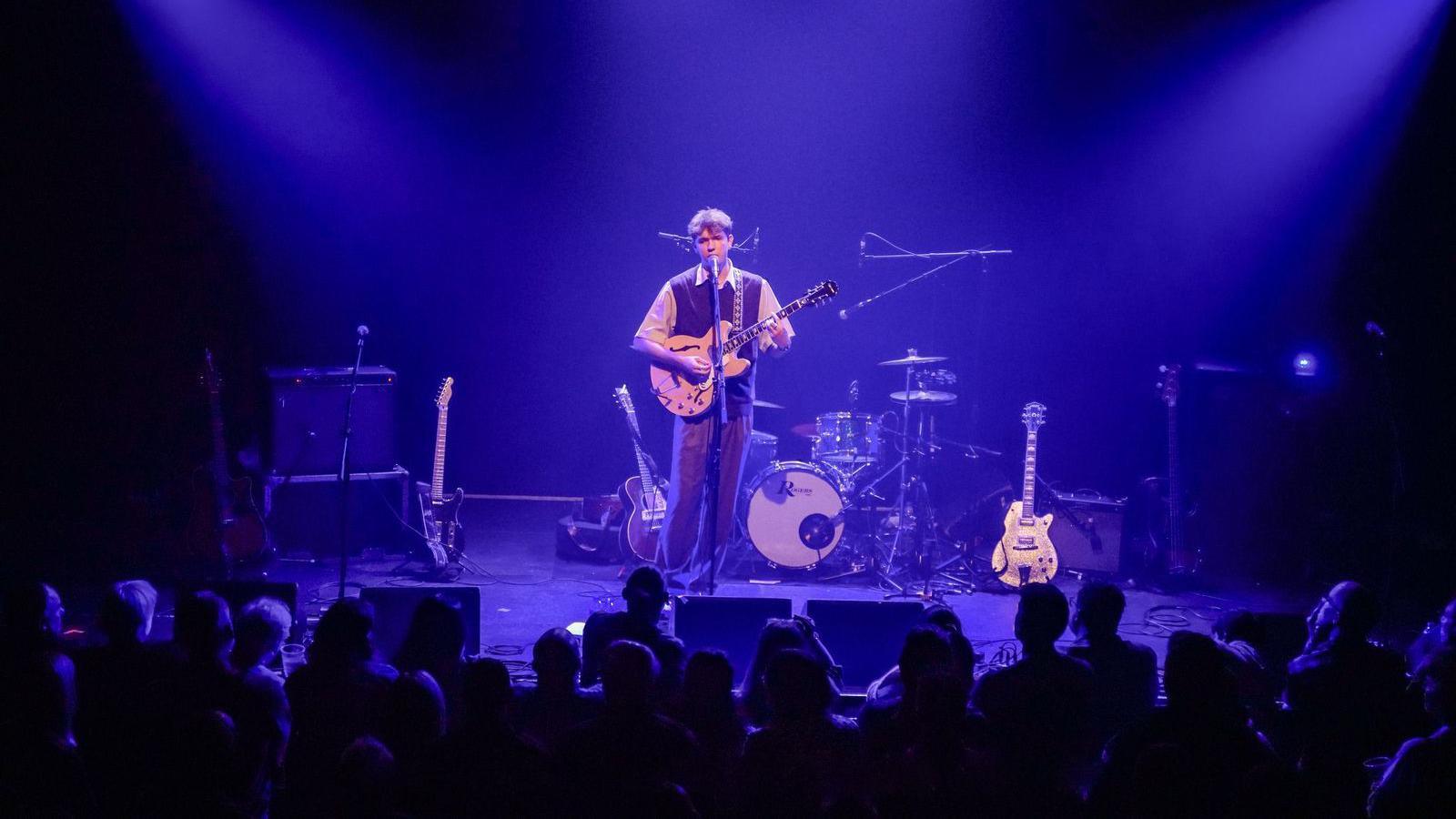 Solo male musician on-stage singing and playing an electric guitar, with blue backlighting