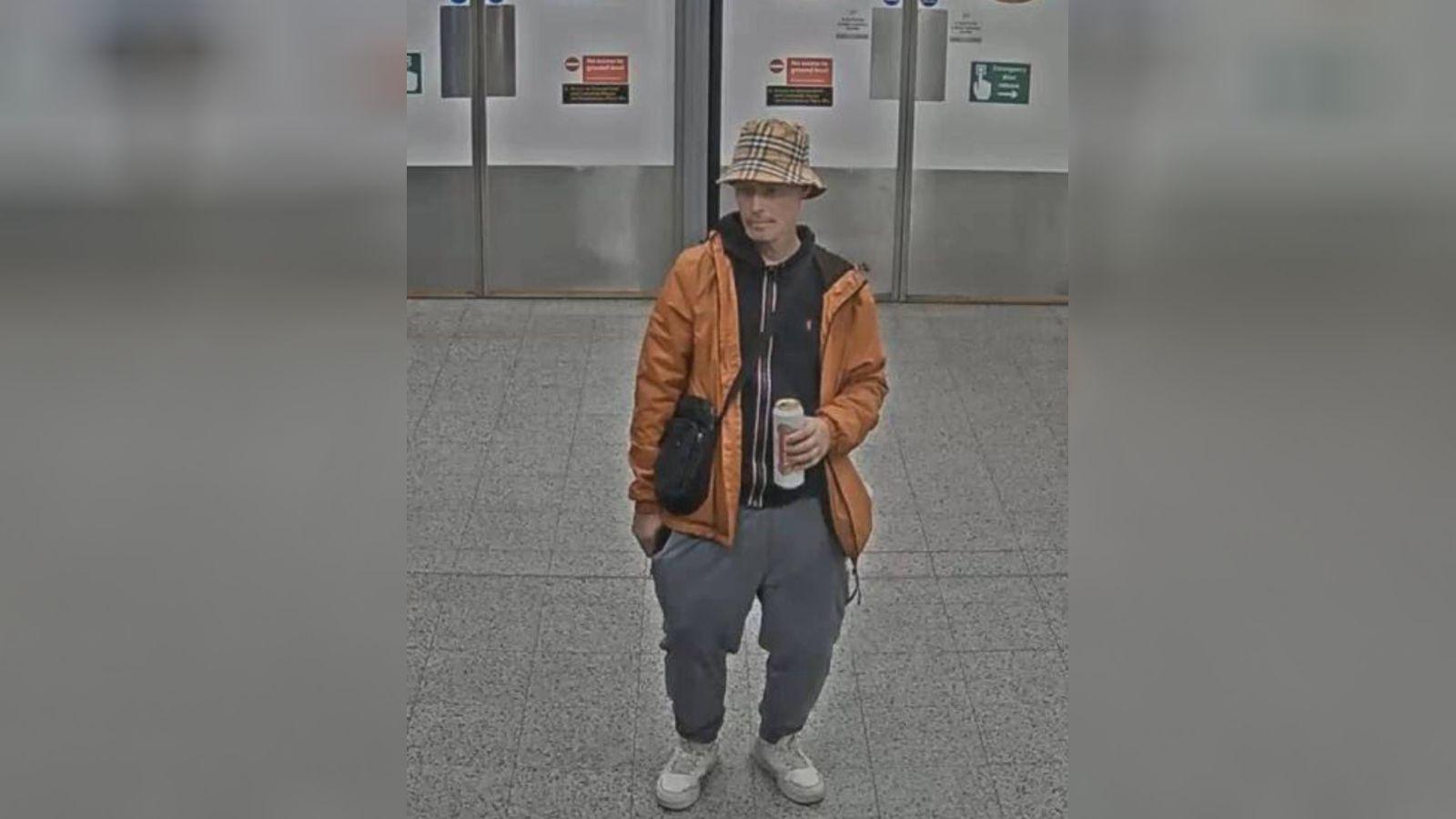 A man in an orange jacket with a bucket hat. He is drinking a pint can of Stella. 