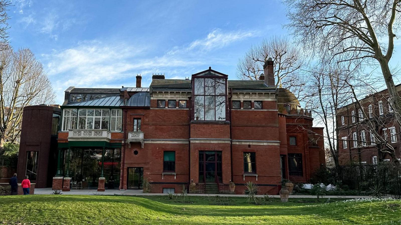 Rear view of Leighton House in London on a clear day, showing its red-brick facade, large glass conservatory, and the distinctive golden dome of the Arab Hall, with two people walking on the lawn in the foreground and leafless trees surrounding the building.