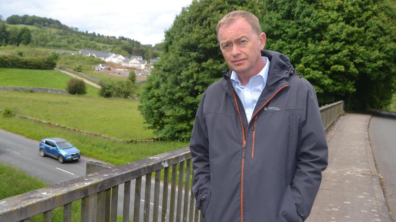 Tim Farron standing by the bridge on Brigsteer Road. He has short fair hair and is wearing a light blue shirt and grey jacket. Below him, two vehicles are being driven along a road. In the background are fields and trees.