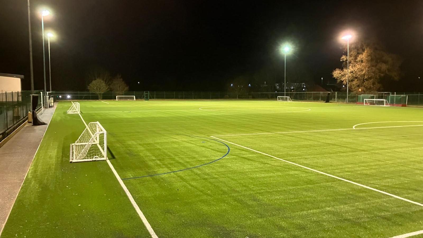 View of brand-new AstroTurf on a football pitch. It is dark and the floodlights are on.