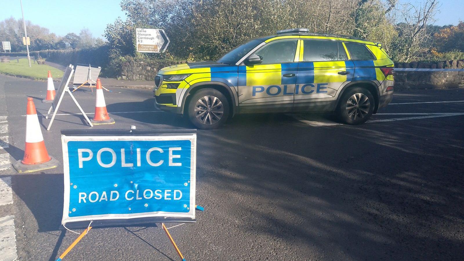 A police car sits at a road block outside the scene that police have cordoned off. There is a sign with 'police, road closed' 