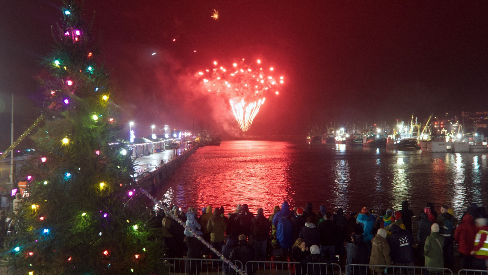 A crowd of people stand next to a Christmas tree as they watch fireworks over a Cornish harbour.