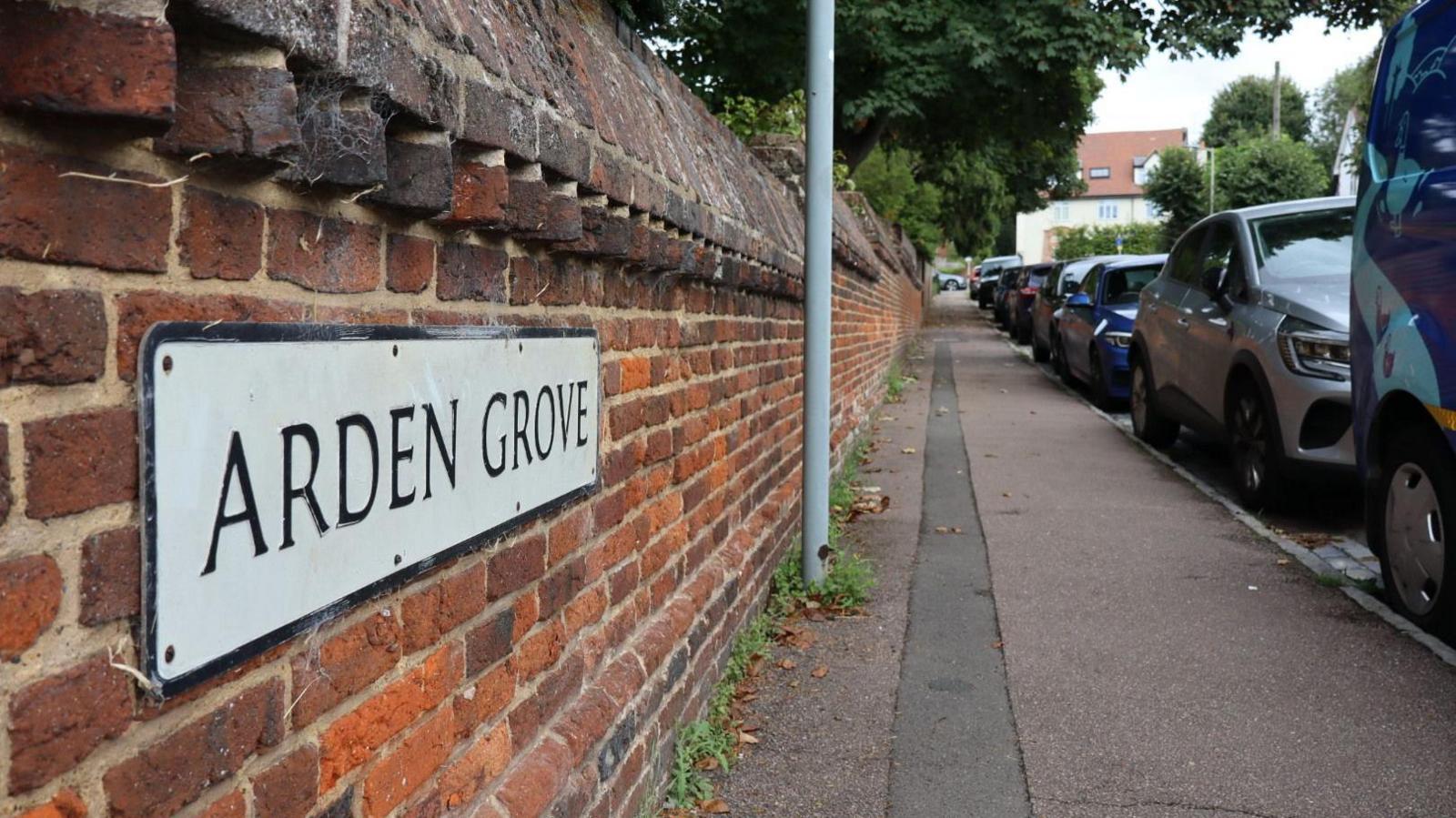 A brick wall with a street sign saying "Arden Grove" next to pavement and a row of parked cars