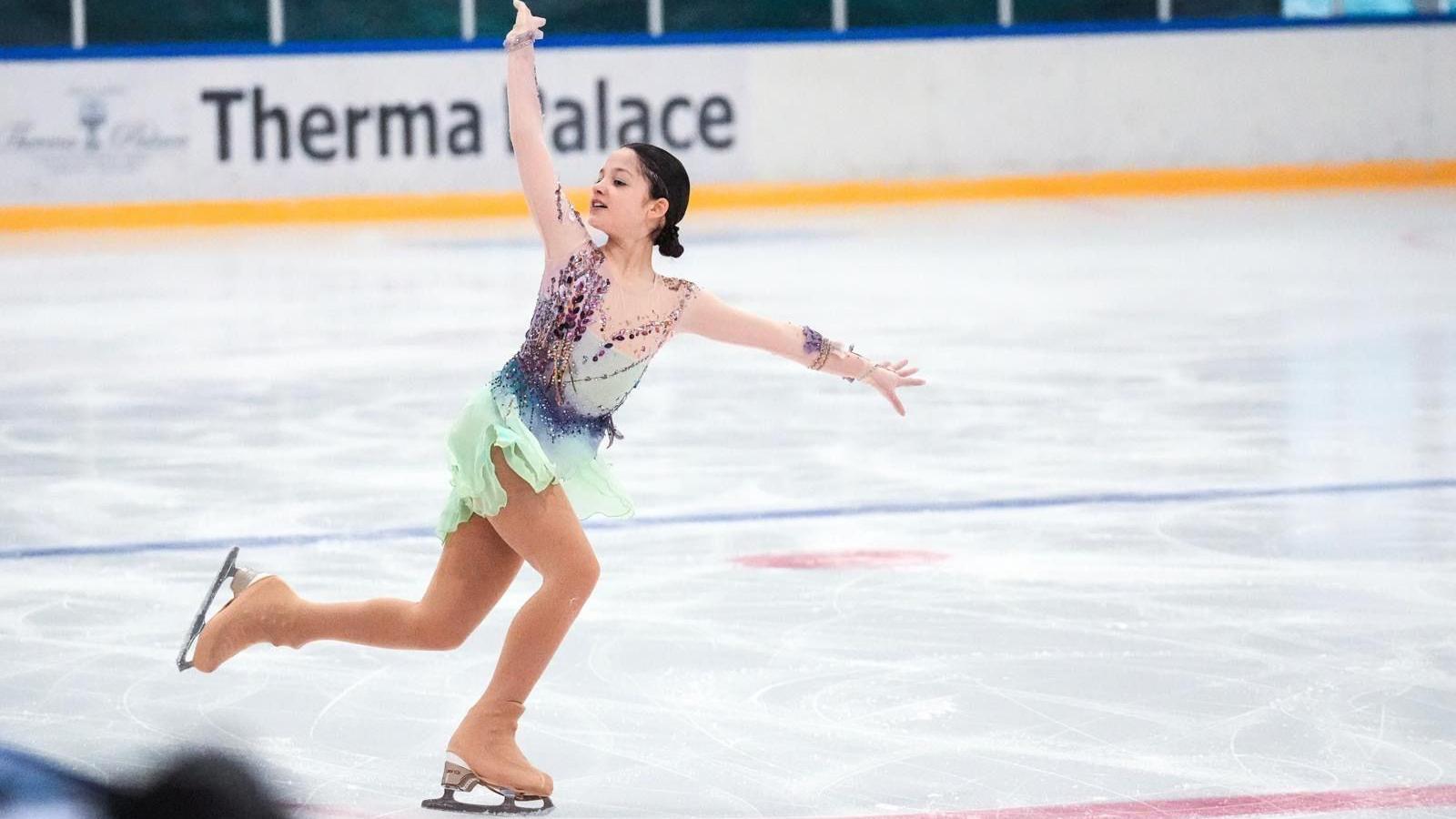 Leilah is figure skating on the ice with her arms outstretched and one leg risen behind her, smiling.