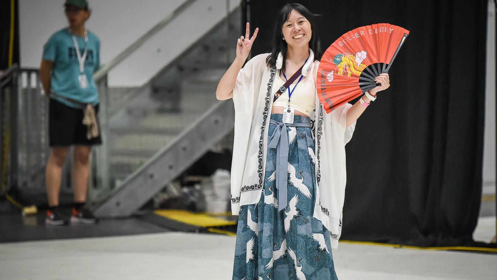 Li Caswell-Sou in traditional hanfu dress, smiling for the camera, while giving a peace sign and holding a red folding far of the Team Chinese Nation roller derby team. A member of the event's staff can be seen behind her.