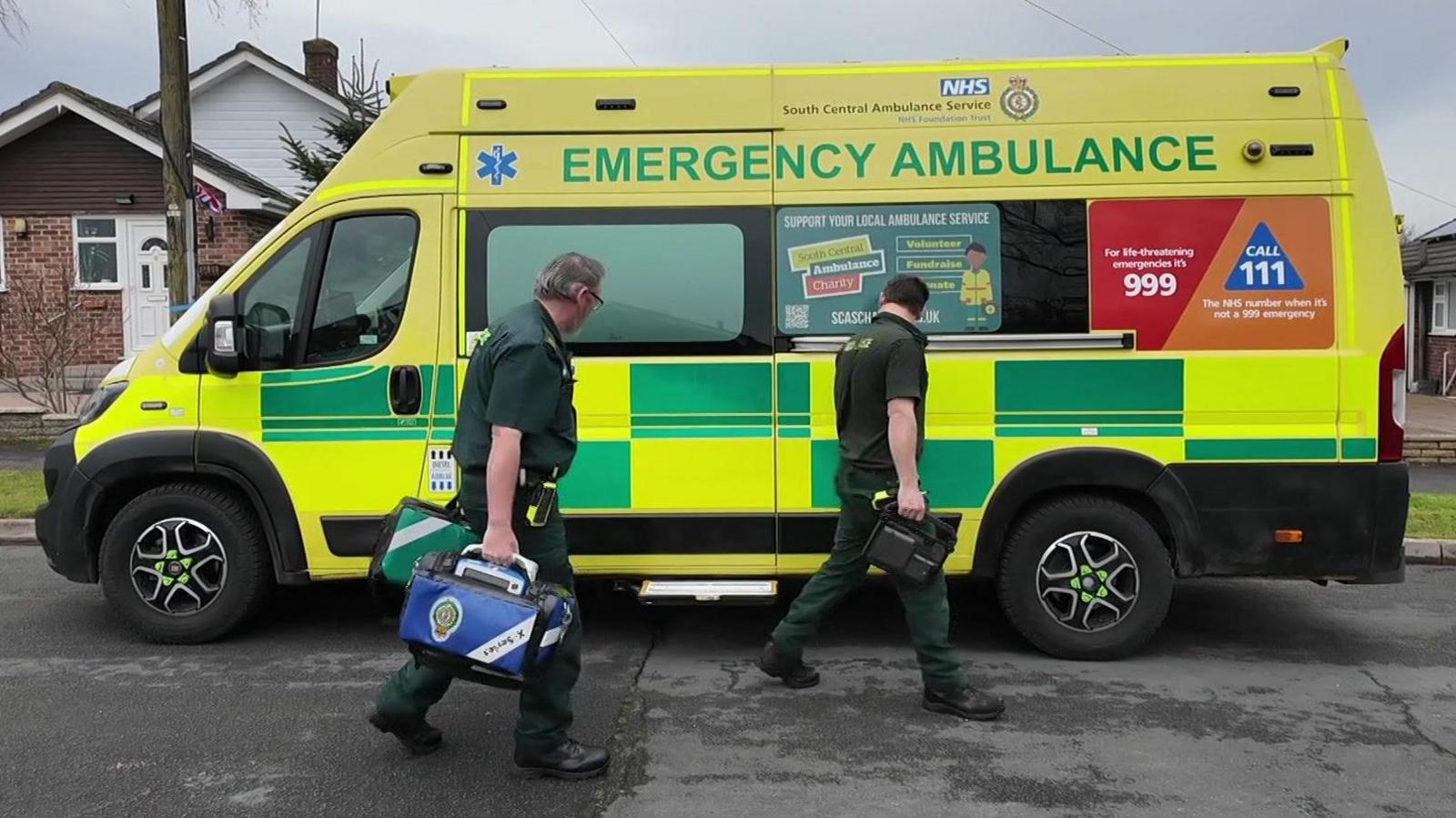 Two male paramedics walking on a residential road outside a yellow and green NHS ambulance. They are wearing green paramedic uniforms and carrying medical bags. In the background is a bungalow. The ambulance has "emergency ambulance" written on it.