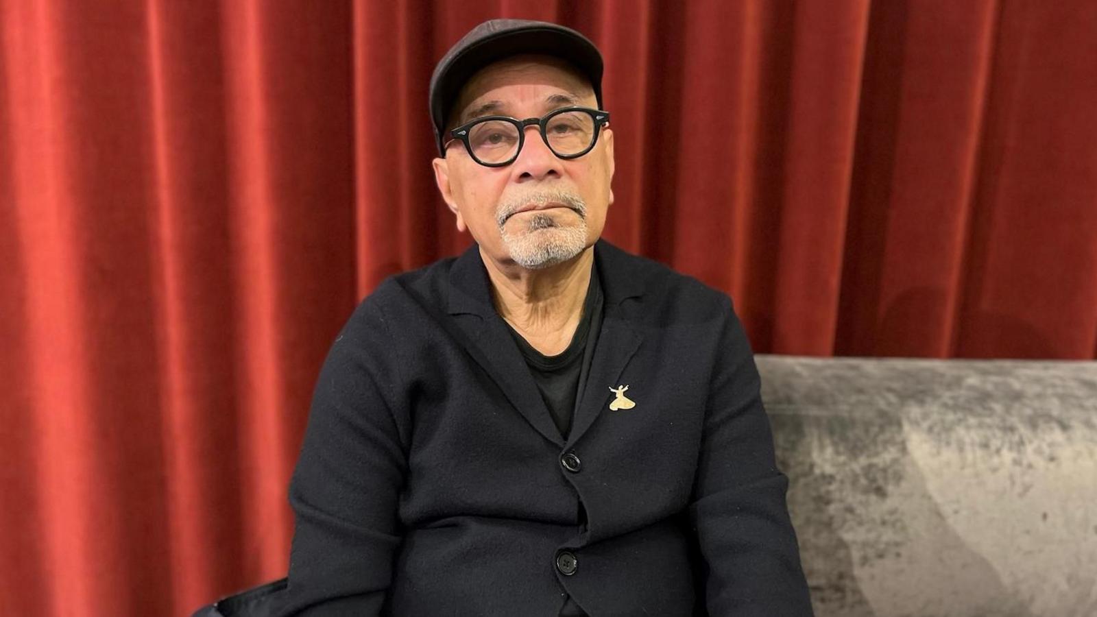 Portrait picture of Ahmed Alauddin Jamal. He sits on a grey velvet sofa wears a grey baseball cap, navy button down jacket, black t-shirt and a golden brooch on his lapel. Behind him is a thick red curtain.