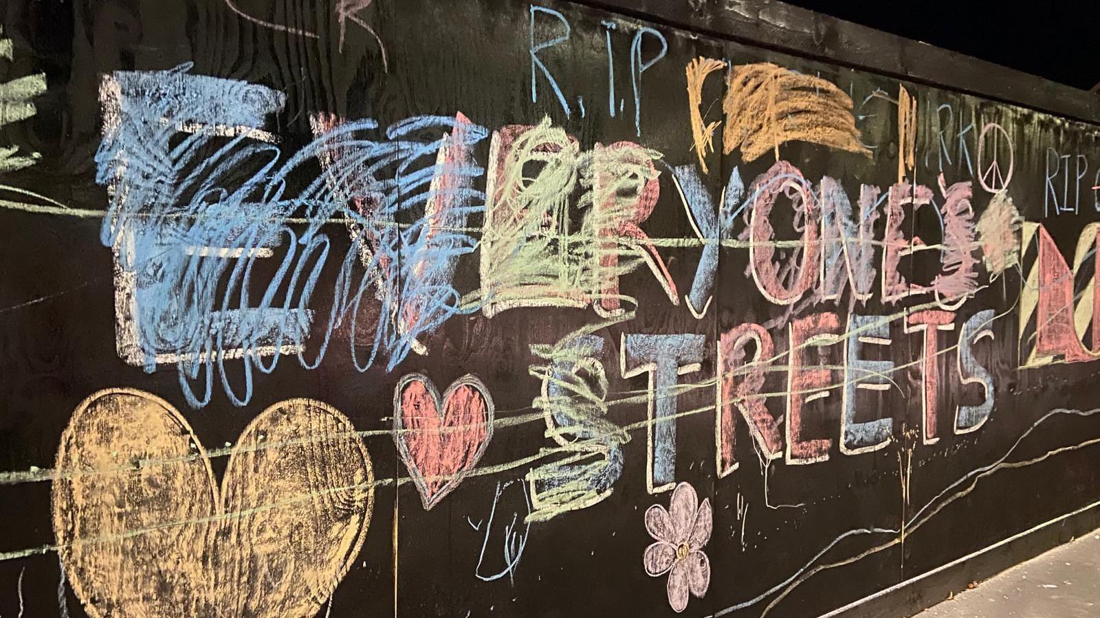 A picture taken at night of a black board along a street that has chalk messages written on it and which have then been later scribbled out. 