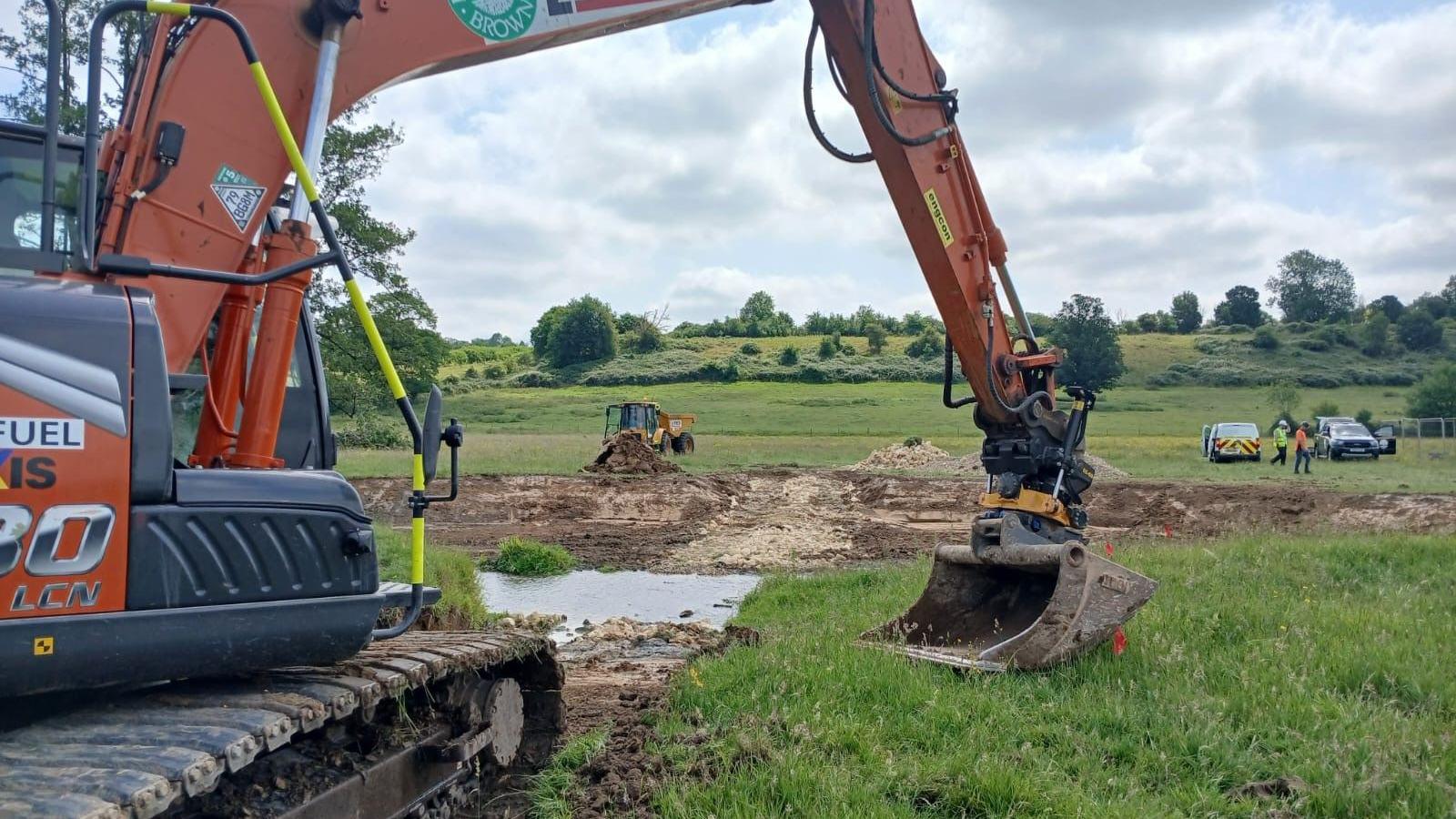 A digger, in a wildlife area, with a river being worked on behind, with three other vehicles, around the site and two people standing by a car. There is grass all around the area. 