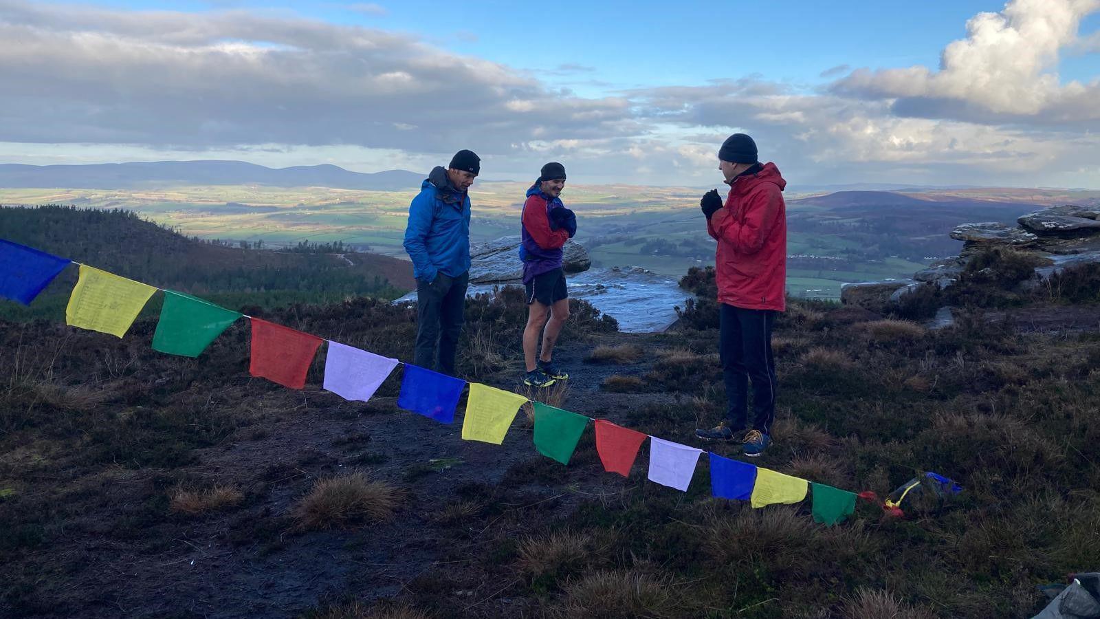 Three men stand at the top of a hill with views for several miles in the distance. It is a sunny but wintery day and they are standing behind some flags which are anchored to a Christmas tree  