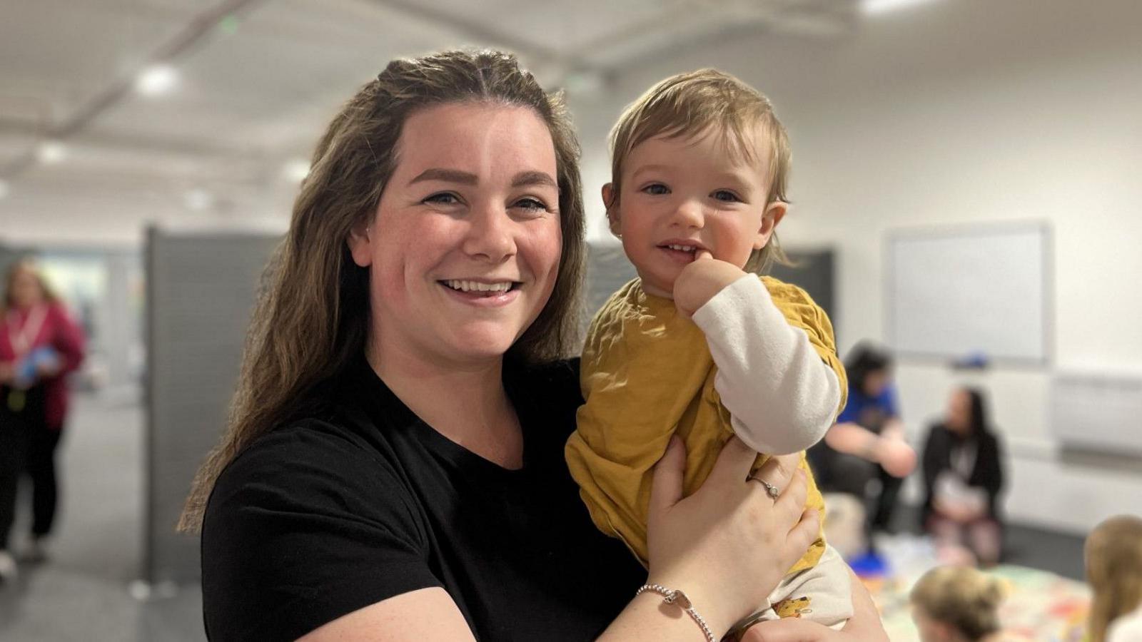 Sarah holding a young toddler. She has long wavy brown hair pulled back off her face and is wearing a black t-shirt and a silver bracelet. The baby has light brown hair and is wearing a mustard coloured top with white sleeves. They are both smiling and the baby has a finger in his mouth. Other women are out of focus sitting in the background. 