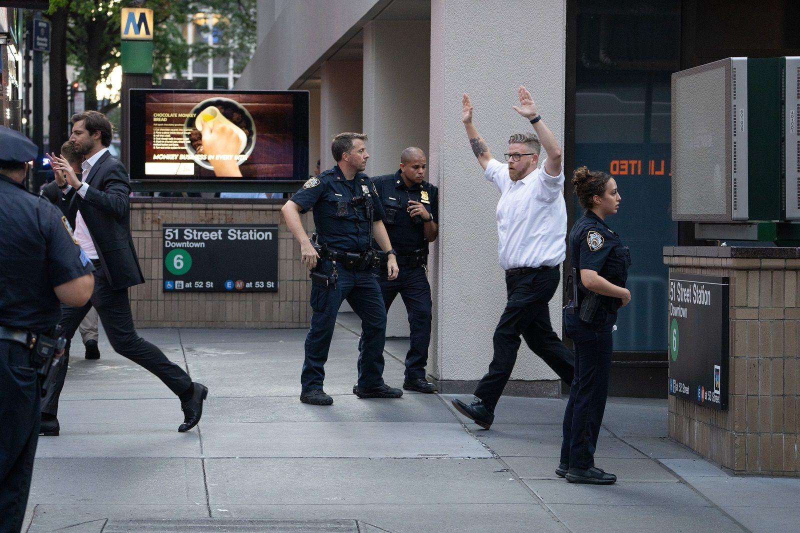 A man leaves a building, walking past police officers, with his hands up. Another man can be seen far left also with his hands up. In the background there is a sign for the New York city subway