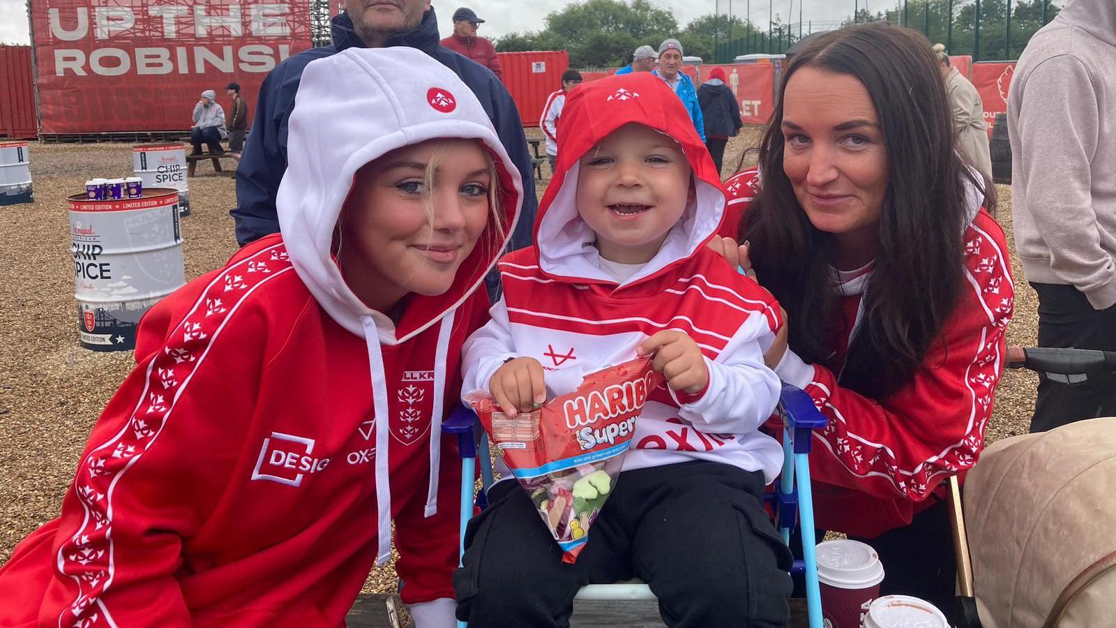 Three Hull KR fans wearing red and white hooded sweatshirts. The woman on the right has long brown hair and is holding on to the back of a deck chair on which a child, who has their hood up, is sitting. The child is in the middle. On the left is a young girl who also has a hood up over her head. All three are smiling at the camera.