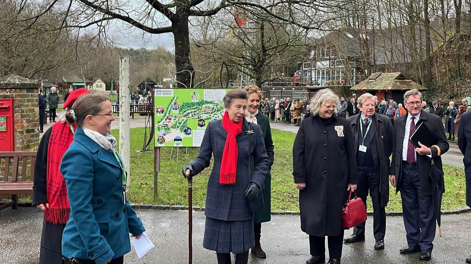 Princess Anne wearing a long, dark blue coat and red scarf. She is holding walking stick in her right hand and is surrounded by half a dozen people. They are on a driveway with green grass and a tree behind them. there is a red post box built into a brick wall to the left. The sky is grey