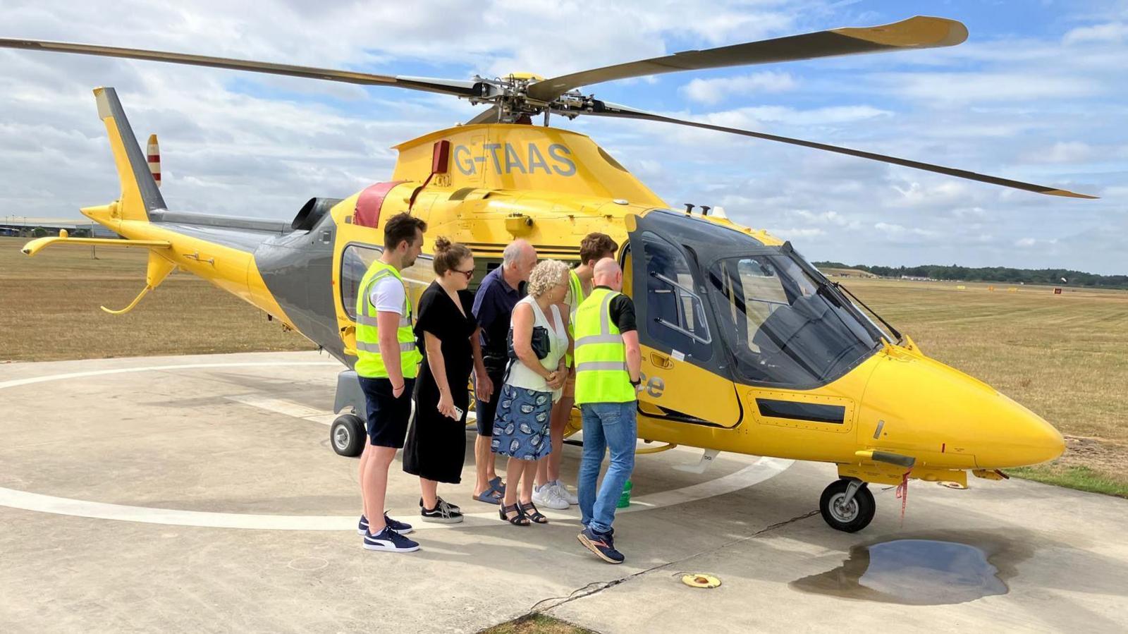 Five people, looking inside an air ambulance. They are all leaning in. The air ambulance is yellow and black, is standing on a concrete ground, with grass in the distance. There are also trees in the distance.