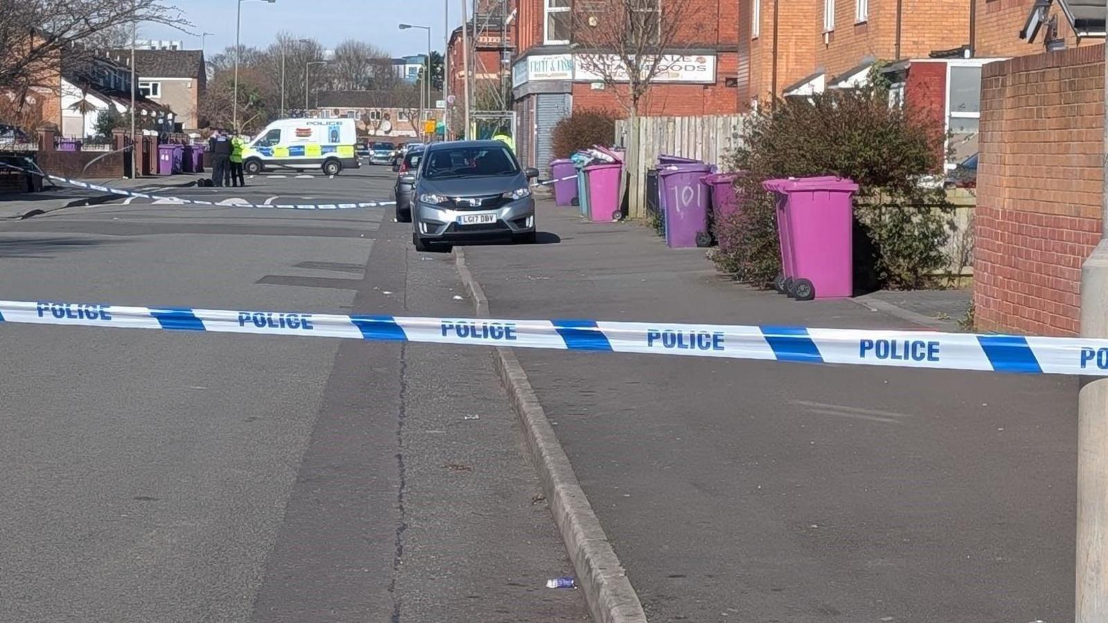 Police tape can be seen in the foreground, tied across a residential street. Pink and purple bins line the side of the pavement. In the background there is a police van and a group of officers are standing in front of it. 