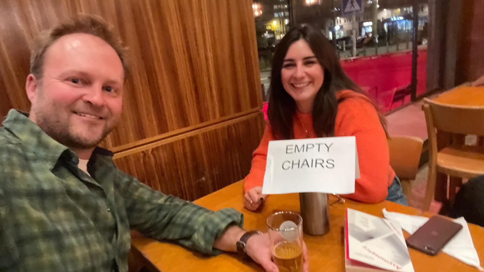A man with a green chequered shirt sitting at a table with a woman in an orange jumper with a little sign saying "empty chairs" on their table