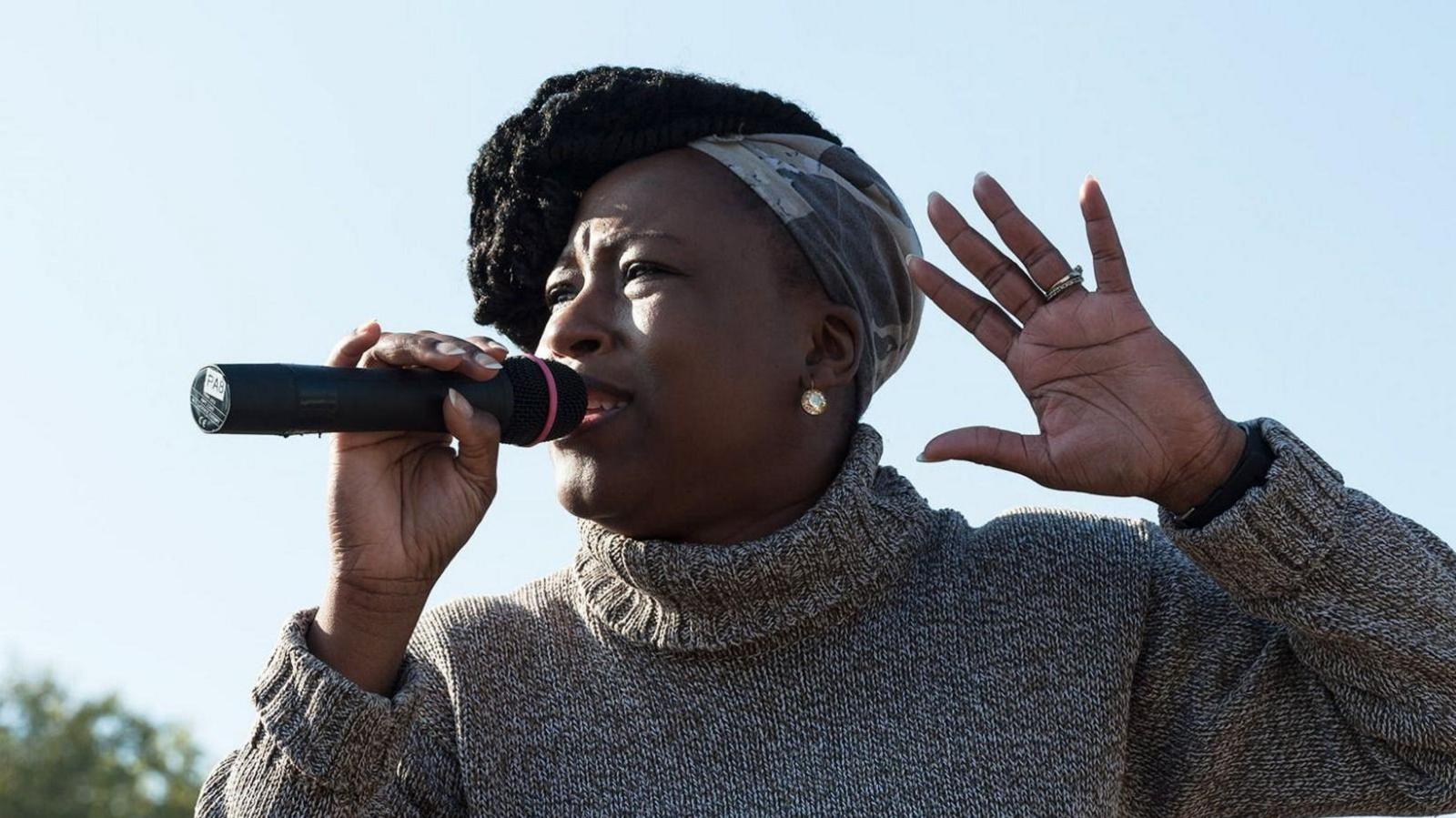 Shola Mos-Shogbamimu photographed outside, against a blue sky, with a hint of a tree in the background in the bottom left hand corner. She is looking off-camera, towards the left of the frame and is holding a microphone in her right hand. She is pictured giving a speech and is mid-sentence. Her other hand is emphasising what is being said - held up to her head height, with her fingers spread and palm showing. She is wearing a rollneck black, white and grey sweater.