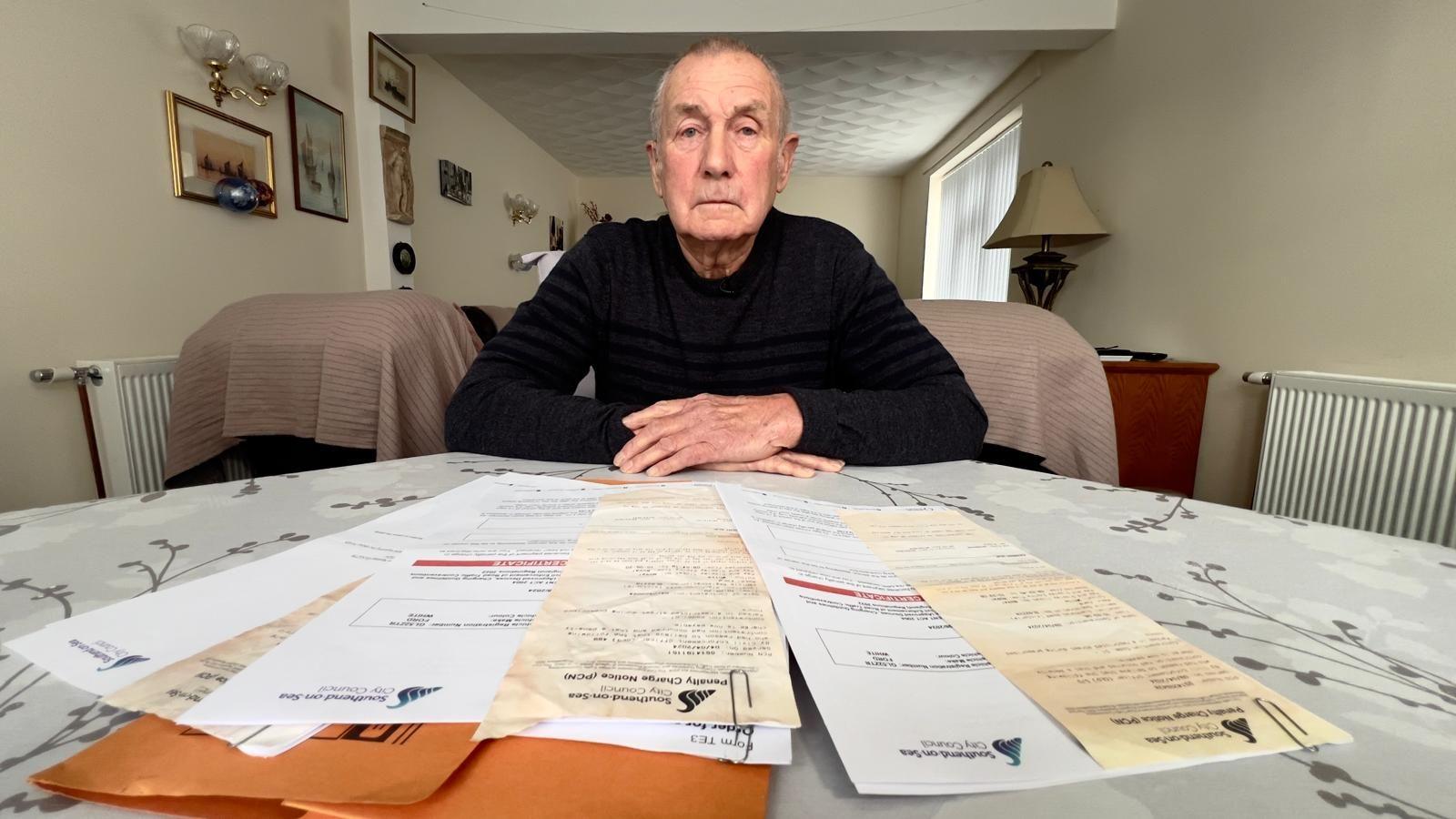 Barry Jager, father of Shaun Jager, sitting in his lounge with several parking fines laid out on a table in front of him. Mr Jager, wearing a black top, has his arms crossed and is looking directly at the camera.