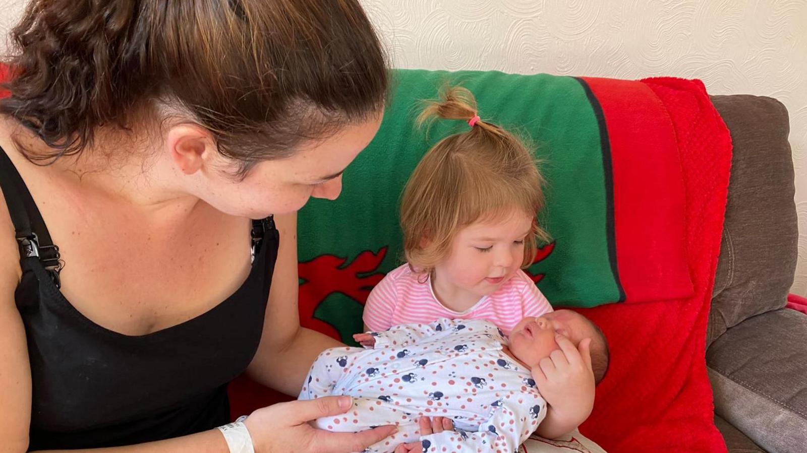 A woman with tied-back dark hair, wearing a black vest top and sat on a sofa with a Welsh flag blanket draped on it. She is looking down at a toddler girl sat beside her, with short fair hair and a pink and white striped t shirt. On the little girl's lap is a newborn baby in a white, blue and pink patterned babygrow. Both the girl and woman have their arms round the baby.