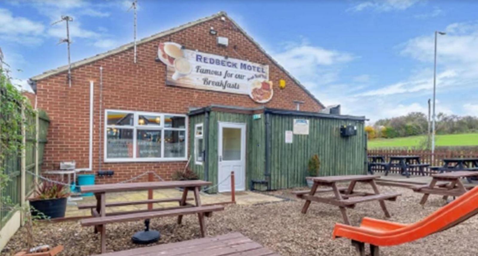 A red brick building with a green extension. A sign on the building reads Redbeck Hotel, Famous for our Breakfasts. In front of the building are wooden picnic benches on gravel. Part of a red children's slide can be seen on the right.