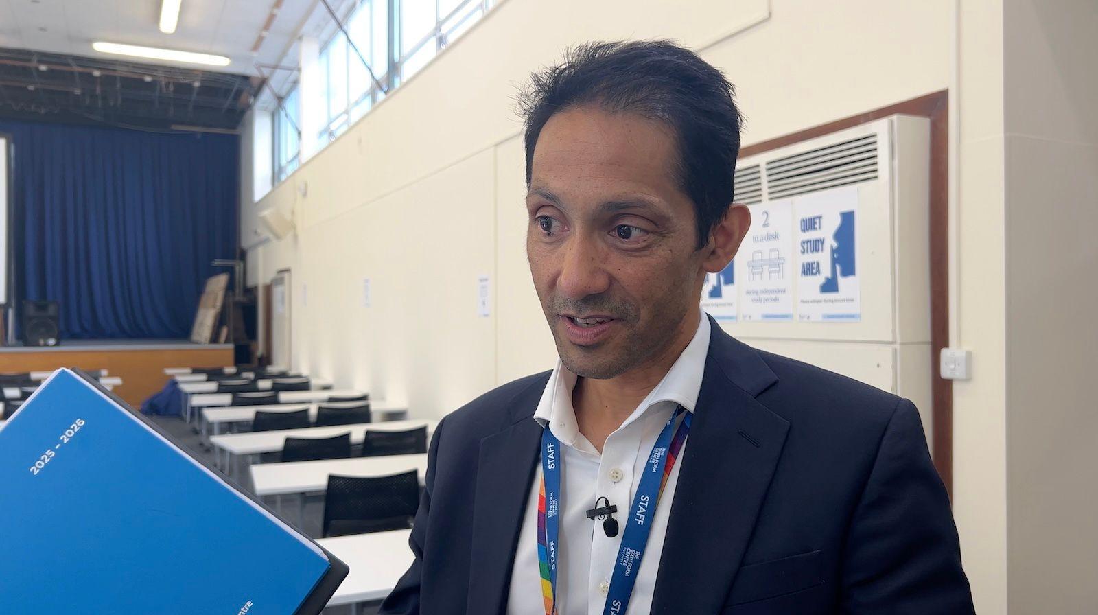 A man in navy jacket and white shirt with the top button undone. He holds a blue academic diary for 2025-2026 and stands in a school hall full of desks.