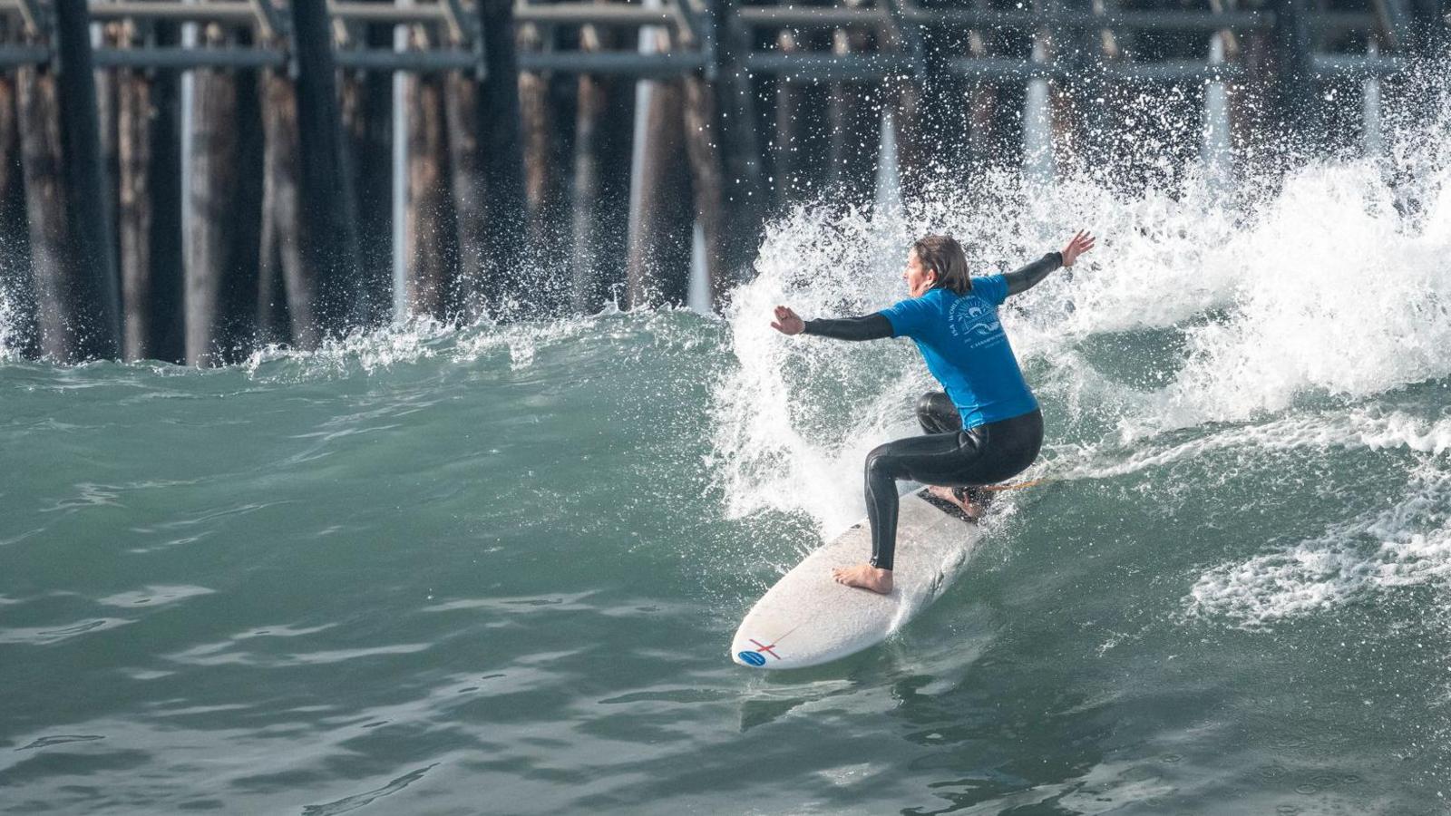 Melissa Reid is surfing a wave with her arms stretched out and her knees bent as the surfboard moves down the face of a wave. She is wearing a Team England T-shirt over her wetsuit. Behind her is the pier.