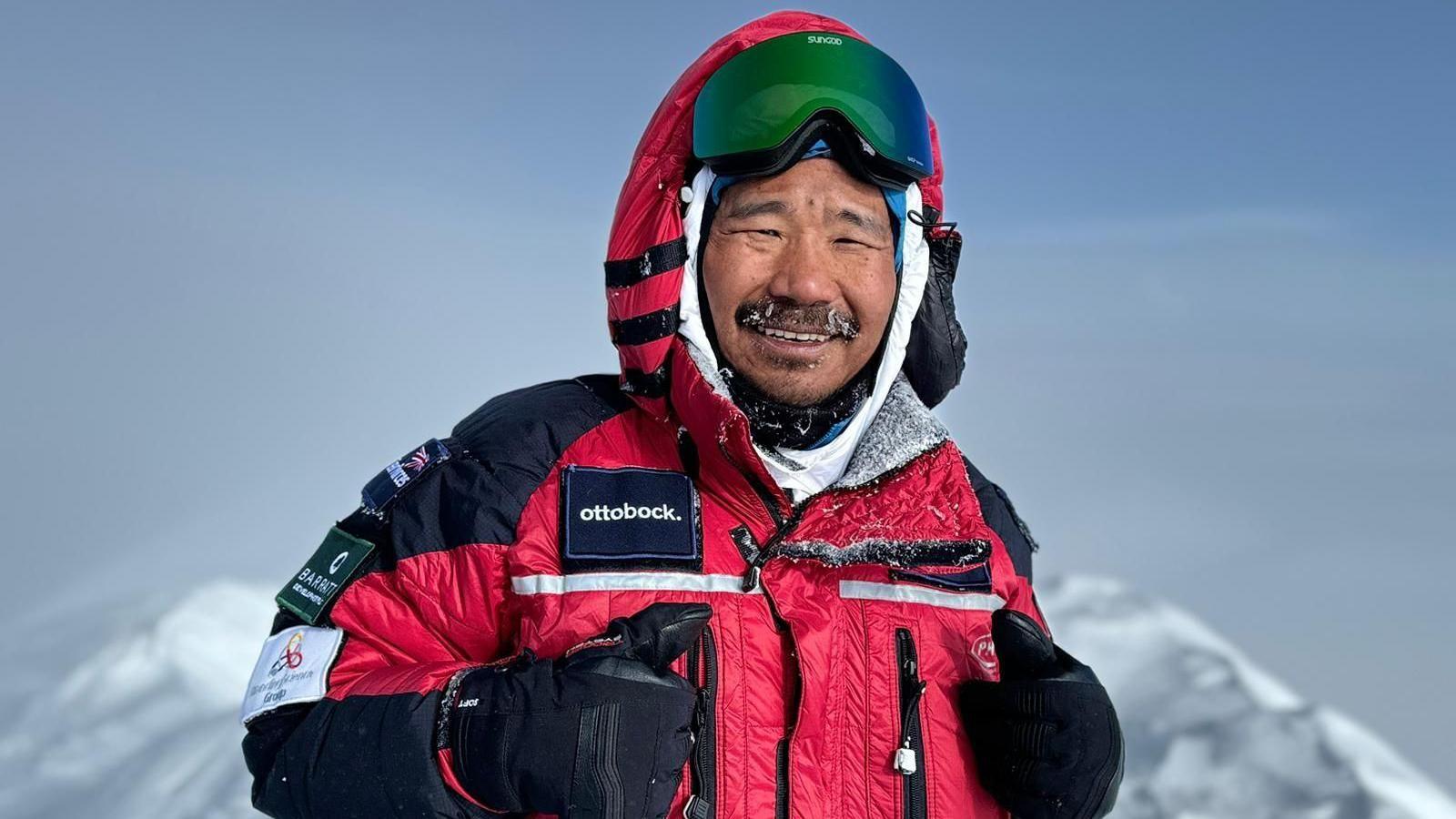 Hari Budha Magar MBE, wearing a red and black coat and smiling at the camera. He is stood atop a mountain with a grey sky in the background.