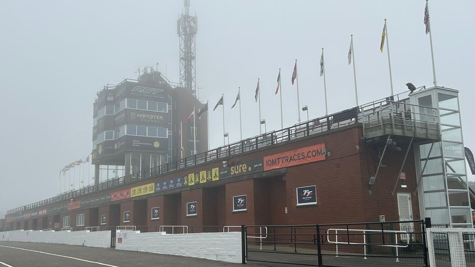 A foggy looking TT grandstand. The red brick building, which has a control tower with large windows around it and international flags along the top.