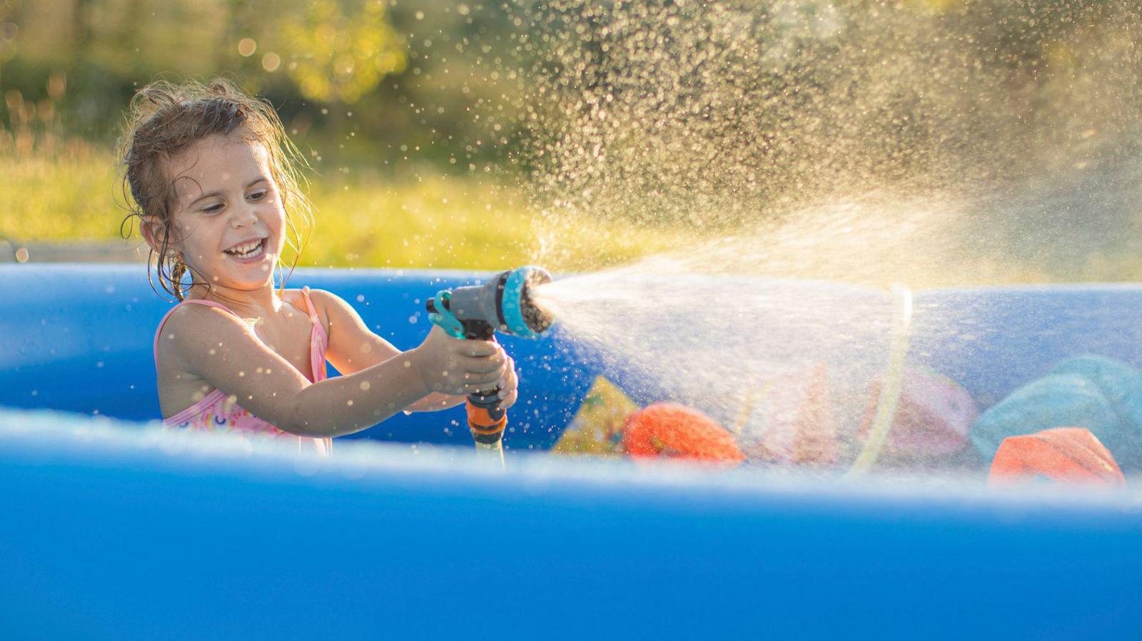 A girl sits in a blue paddling pool spraying a hose pipe in the sunshine