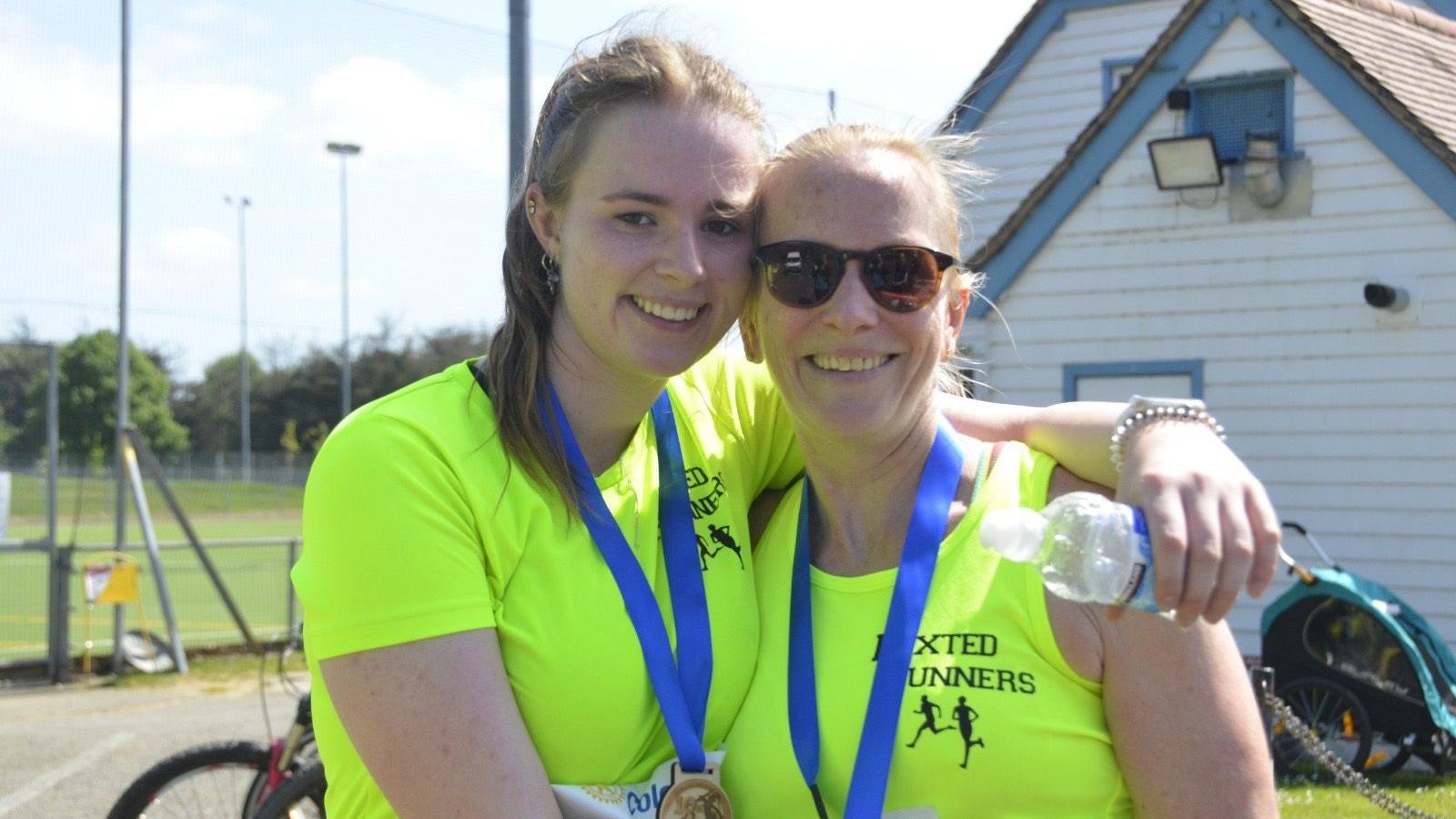 Jamilla Blake with her mother Jan Wilcox at a running track. They are both wearing neon green t-shirts with a running club logo. They are both wearing medals with blue ribbons. They are both smiling at the camera. Jamilla has her left arm around her mum and is holding a water bottle. Her mother is wearing dark glasses. 