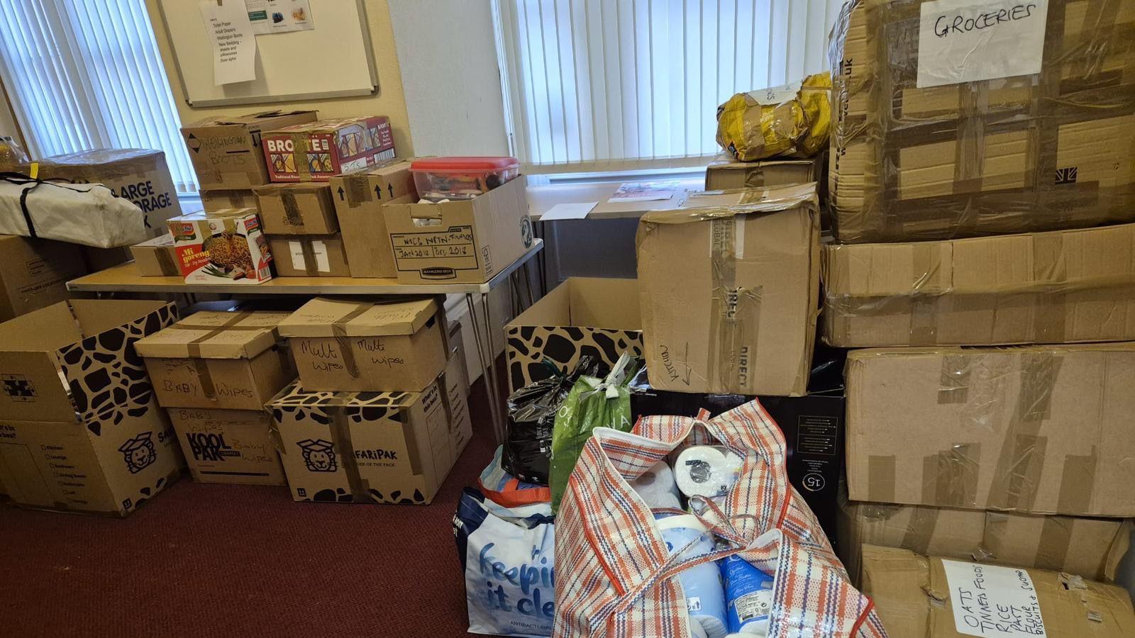 A large collection of donated items, in a room, all piled up. Including several bags of items, with toilet rolls in them. There are two windows, and a red floor.