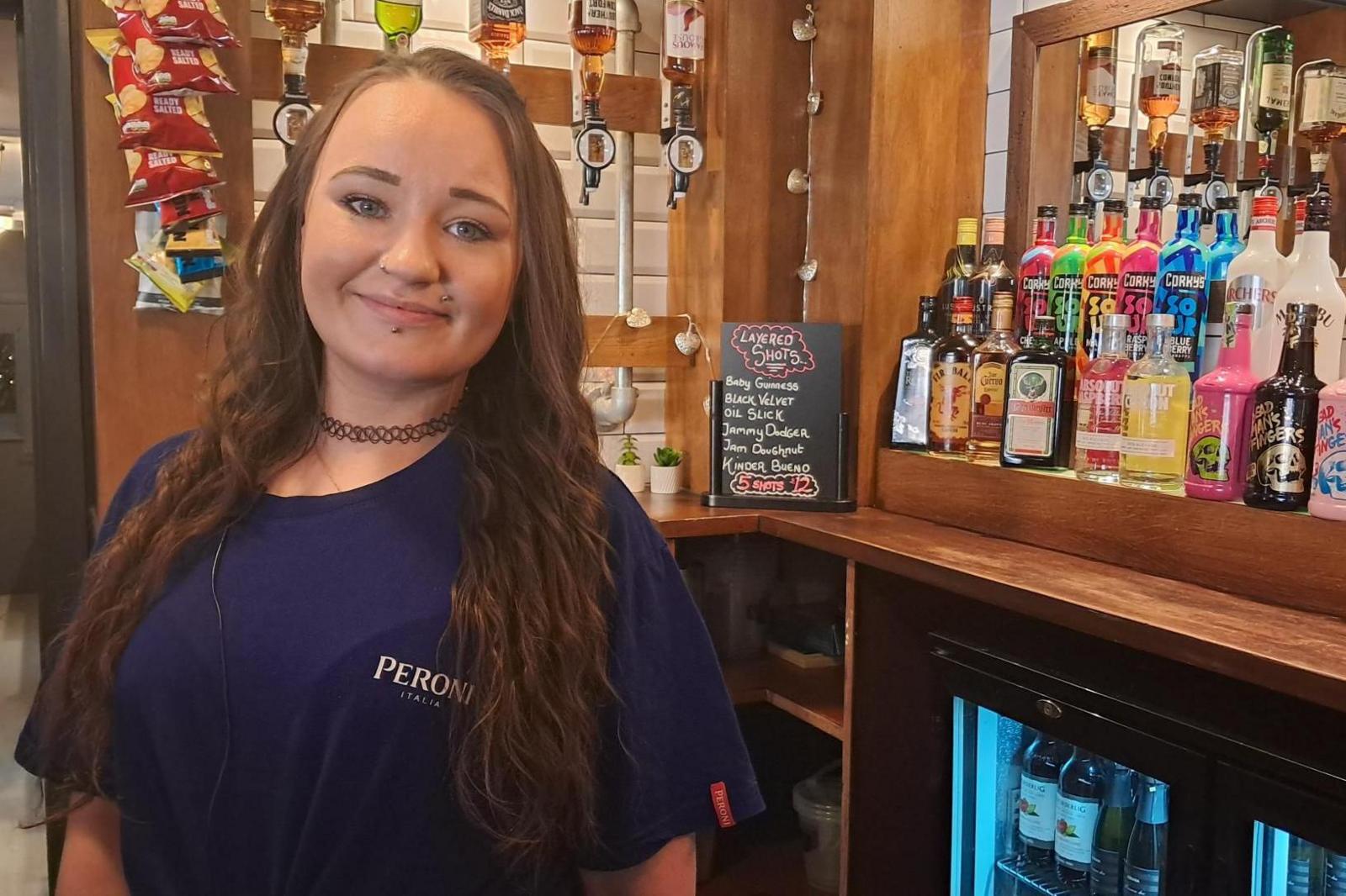 April Smith: a smiling young woman, with long dark hair, stands behind a bar. There are bottles of spirits in the background, and a drinks' fridge pictured bottom right.