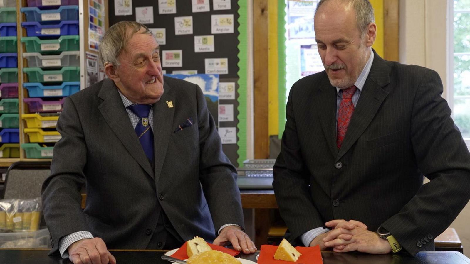 Dr Neville Brown is seated (left) in a smart grey suit while smiling at his son - Dr Daryl Brown (right) in a pinstripe black suit with a red tie, with a slice of cake in front of both of them.