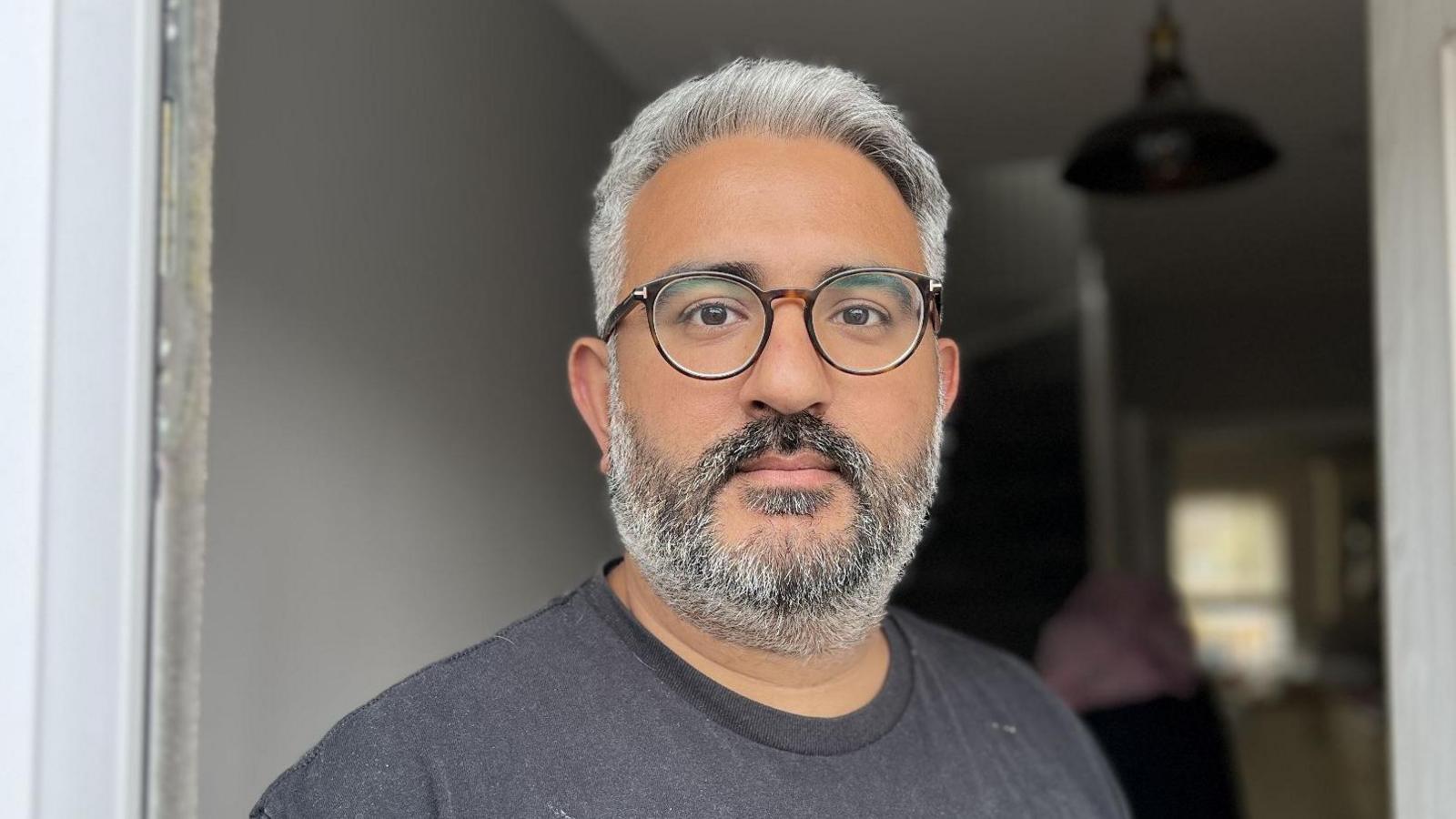 Aman is stood in the centre of the frame, he is wearing round glasses and has grey hair with a grey beard and a grey t-shirt