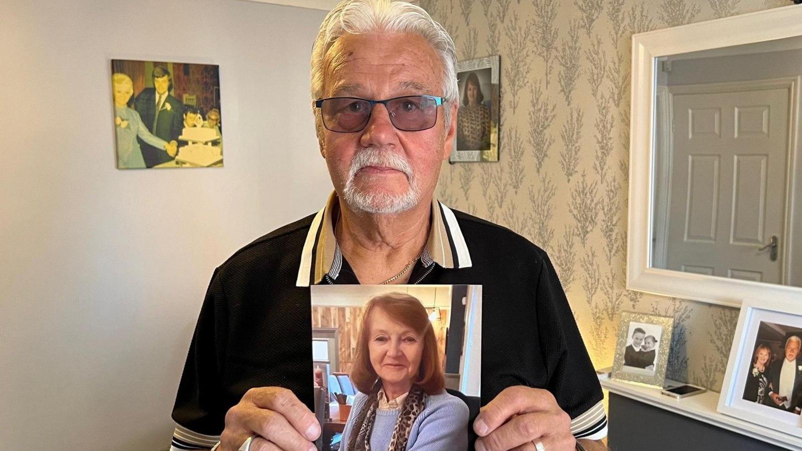 A man in his 70s is stood in the middle of a living room holding up a picture of his deceased wife. The man has grey hair and a beard and is wearing glasses. The picture of his wife shows a lady in her 70s with shoulder-length brown hair. There are pictures of the couple on the wall and on the mantelpiece.