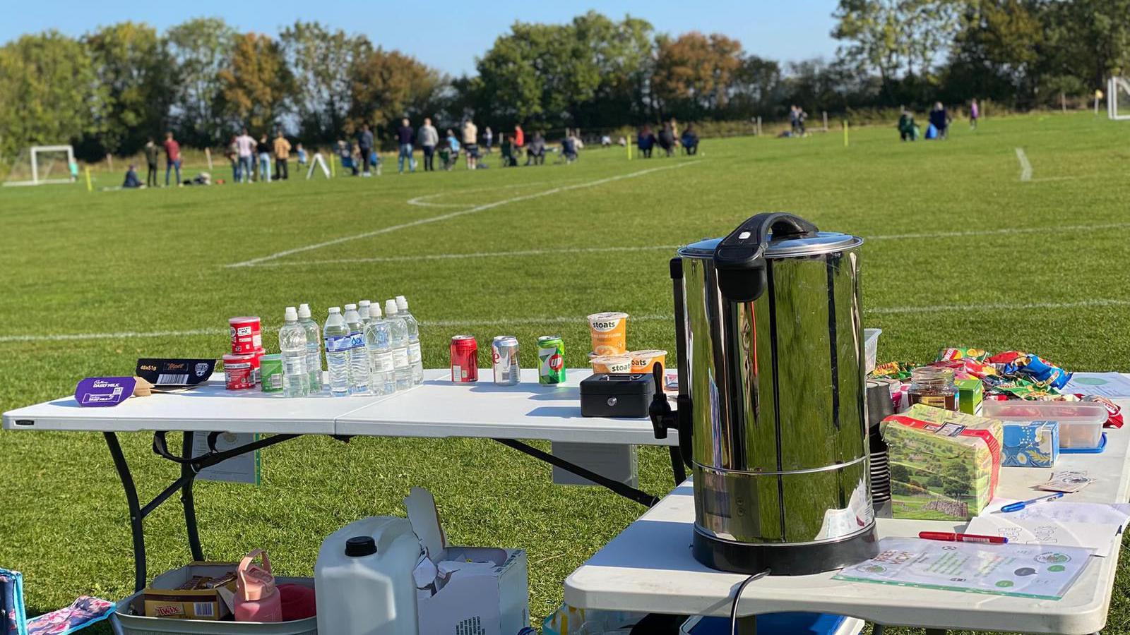 Two stall tables set up at the edge of a football pitch. Bottles of waters, soft drink cans, a steel hot drink dispense, tea, coffee and sweets rest on top of the tables. People on the football pitch and spectators can be seen in the distance.