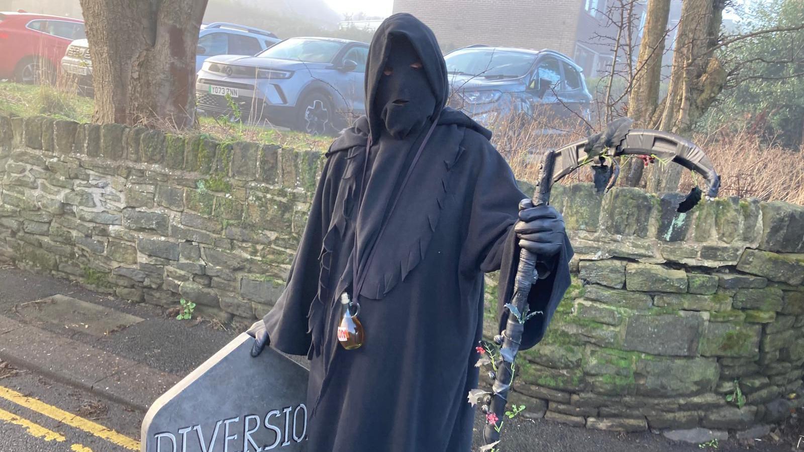 A man dressed as the Grim Reaper holding a scythe and a sign in the shape of a gravestone, which reads "diversion".